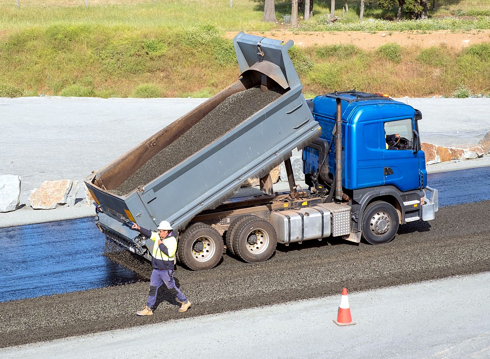 Aggregate being spread onto fresh bitumen while a crew member guides the truck